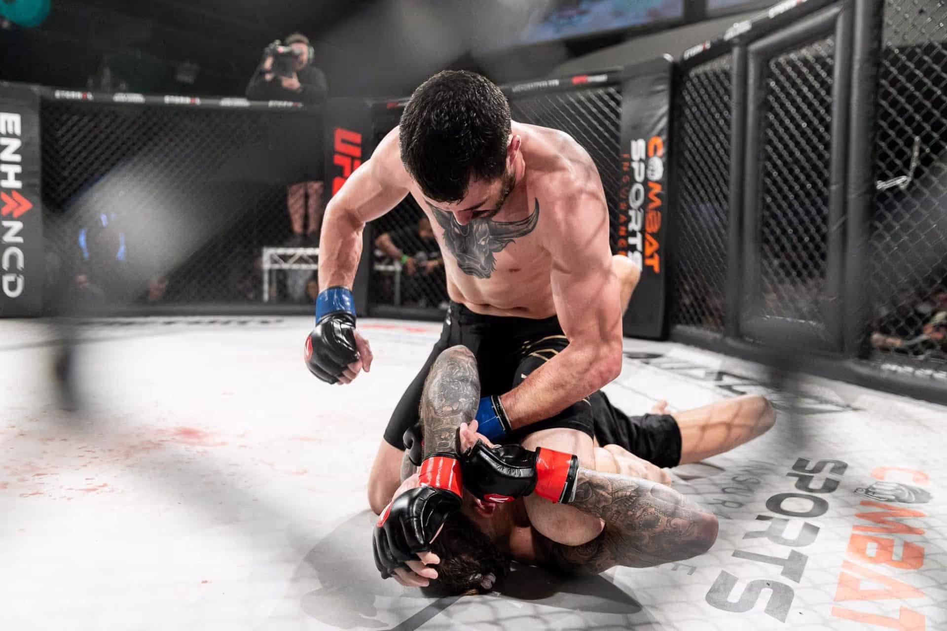 Ground fighting inside the cage at a combat sports event, Combat Sports Insurance branding on the cage padding